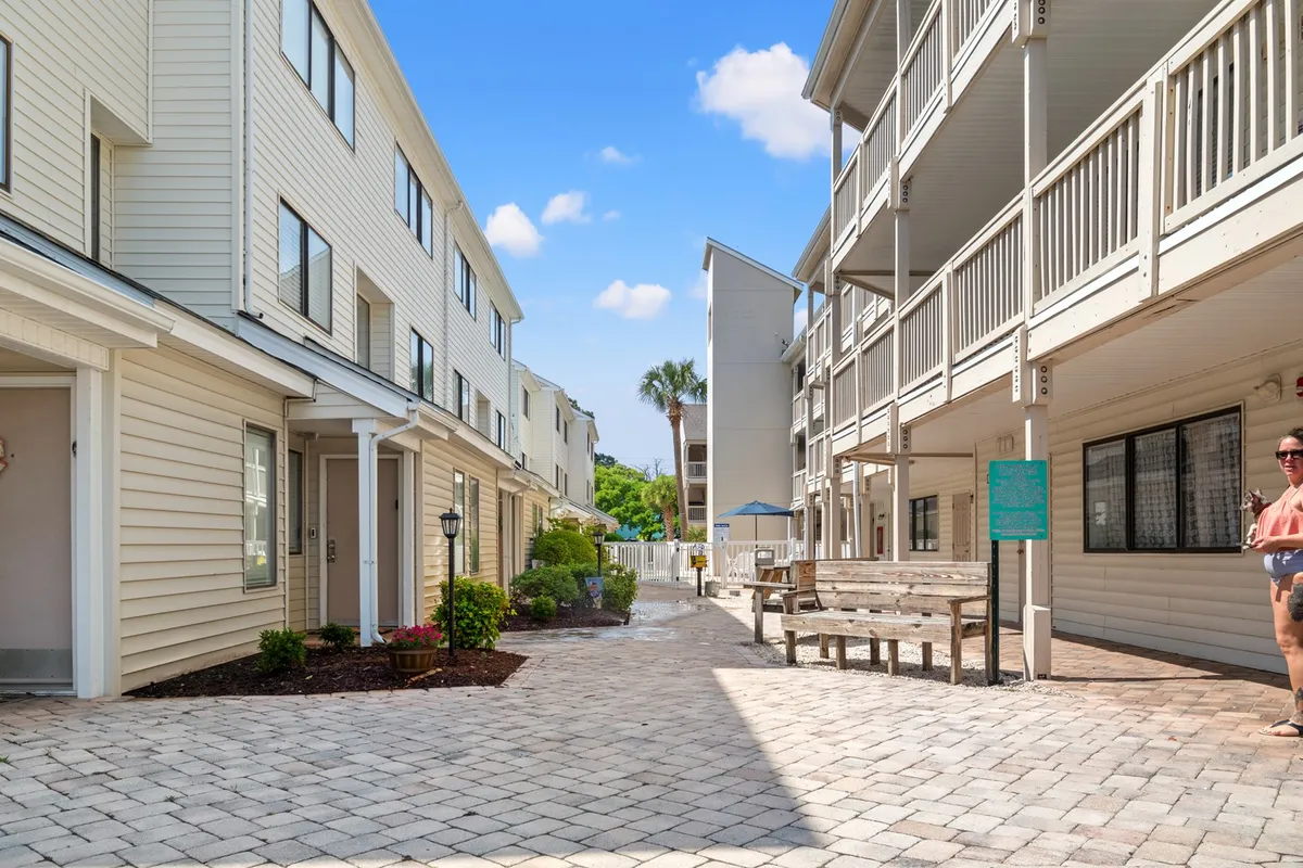 A landscaped garden with paved pathways, benches for sitting, and nearby residential buildings. The setting is calm and inviting, ideal for leisurely walks, socializing, or simply enjoying the outdoors. The benches and flat pathways suggest accessibility and comfort for seniors and snowbirds.