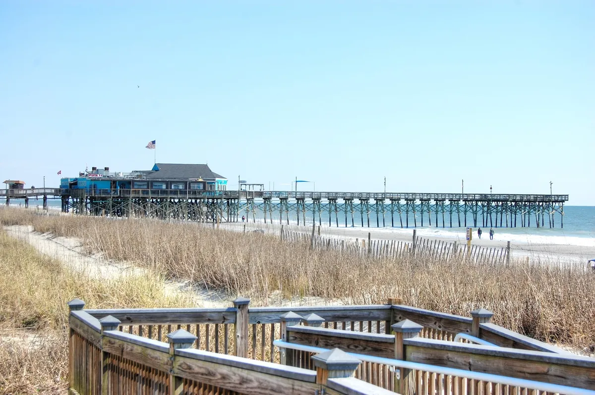A serene exterior view of the beach with a pier stretching into the ocean, surrounded by dune grass and beneath a clear blue sky. The scene is peaceful, inviting guests to walk, fish, or simply enjoy the coastal atmosphere.
