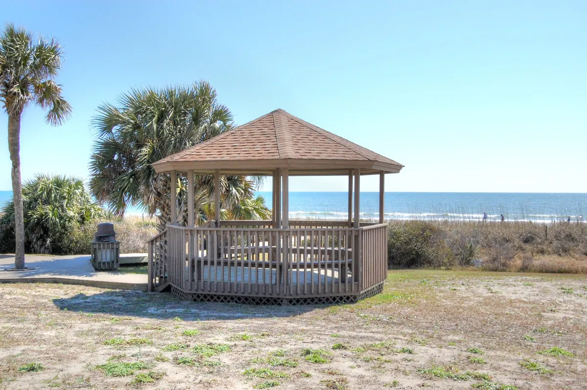 A wooden gazebo sits amidst palm trees with a stunning ocean view in the background. The scene is serene and inviting, perfect for couples or adult groups seeking a peaceful atmosphere. The area is designed for gatherings and relaxation, making it ideal for romantic picnics or quiet moments together.