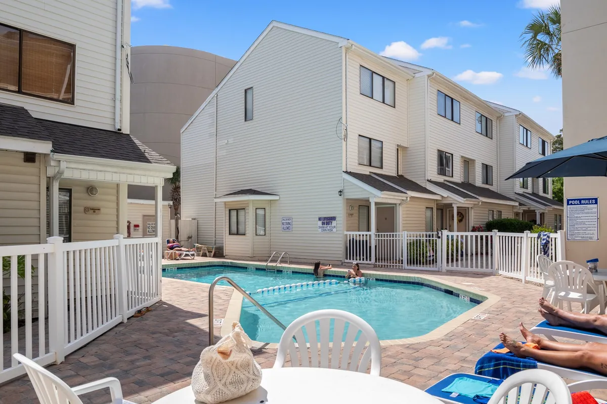 A sparkling swimming pool takes center stage, surrounded by inviting poolside chairs and swaying palm trees, all set against the backdrop of the Ocean Dunes Villas' charming exterior. The scene is bathed in golden sunset light, evoking a sense of relaxation and vacation bliss. The setting is perfect for swimming, sunbathing, or gathering with friends and family for leisurely afternoons and unforgettable evenings.