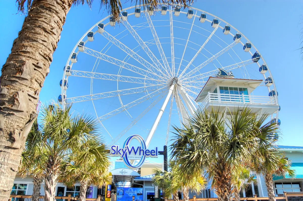 A lively exterior scene showing the iconic Myrtle Beach SkyWheel, palm trees, and a ticket booth bustling with activity. The bright sky and people enjoying their day evoke a sense of excitement and family fun.