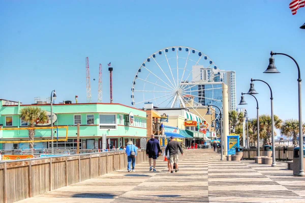 A vibrant exterior scene featuring the Myrtle Beach boardwalk, with a prominent Ferris wheel, colorful storefronts, and people strolling along. The energy is joyful and social, capturing a classic beachside adventure atmosphere.
