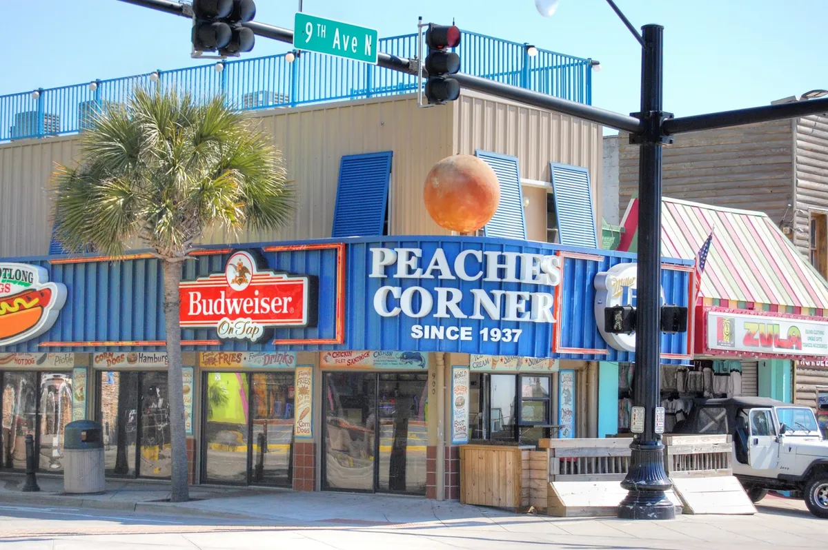 A lively Myrtle Beach street scene featuring the iconic Peaches Corner sign, colorful awnings, and a palm tree. The area is bustling with people socializing and enjoying local cuisine, representing the vibrant entertainment and dining scene nearby.