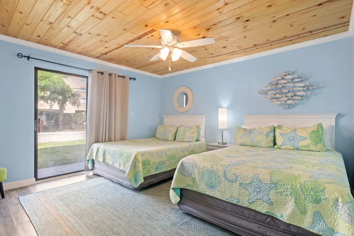 A bright, cheerful bedroom featuring two double beds with colorful bedding, a large window that lets in ample natural light, and a wooden ceiling. The inviting atmosphere suggests comfort and space for multiple guests to relax and recharge.